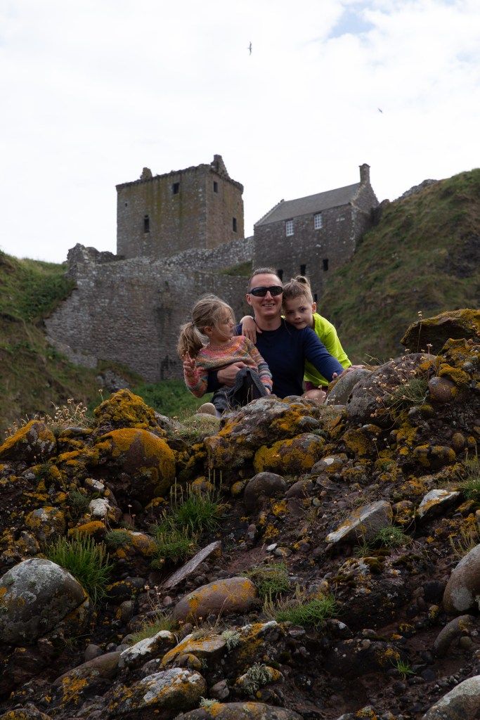 A man and two small children on a stone with a Scottish castle ruin in the background.